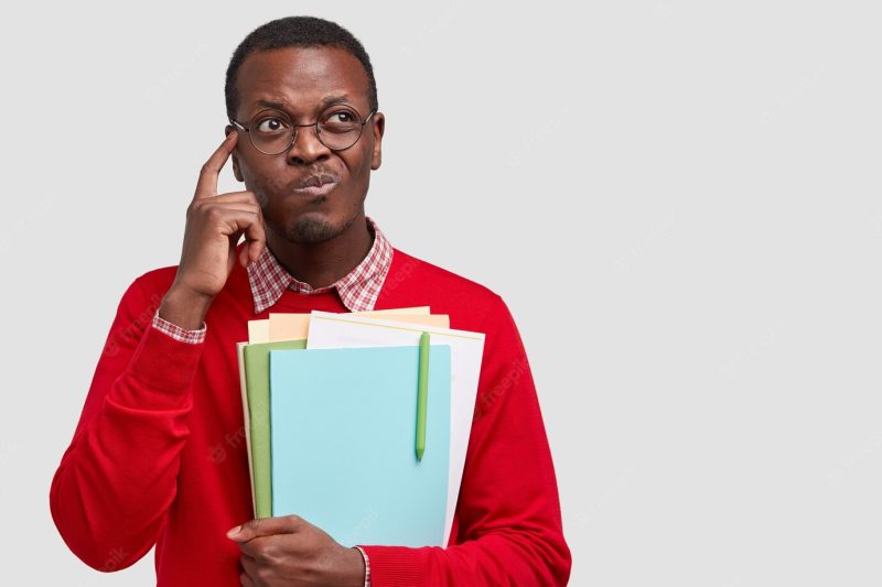 Thoughtful Afro American man purses lips touches temple, contemplates about something, carries folders with pen Free Photo