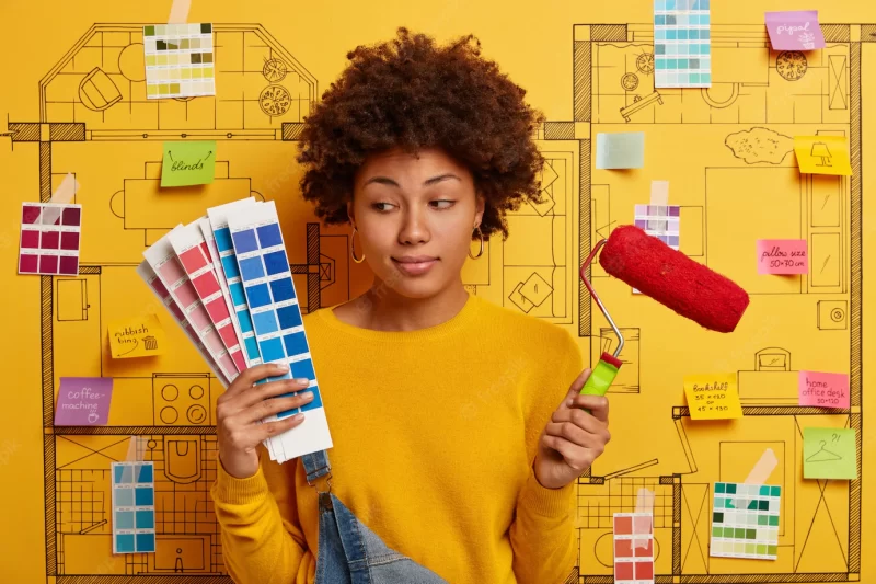 Thoughtful indecisive woman looks at color samples, holds paint roller, thinks about refurbishment of walls in new house, poses against sketch with sticky written notes. repair, building, home concept Free Photo