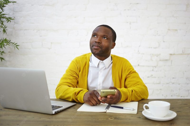 Thoughtful pensive african male freelancer in stylish clothes looking up with thoughtful expression, using smart phone, calculating finances, working at home office, sitting in front of open laptop Free Photo