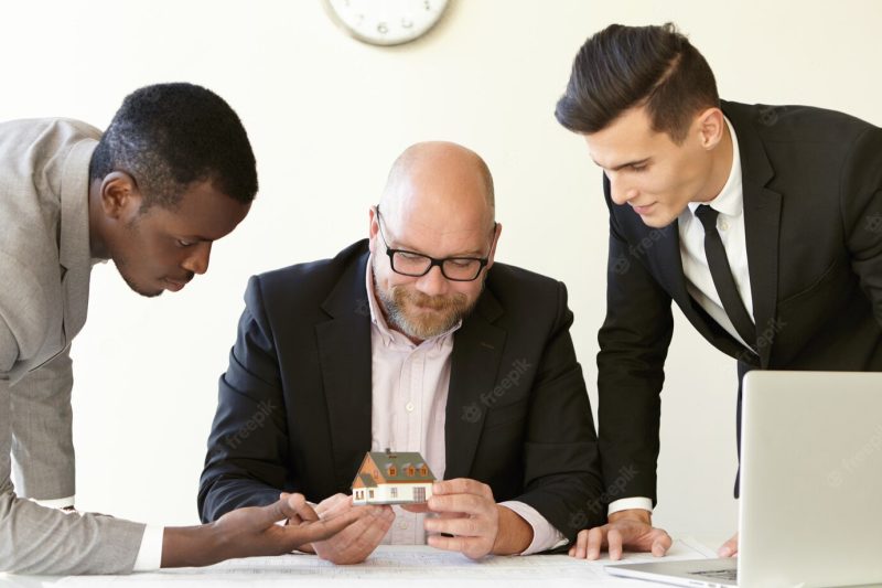 Three office men estimating mock-up model of future terraced house. caucasian engineer in glasses holding miniature and smiling. other colleagues in suits looking at tiny house with interest. Free Photo