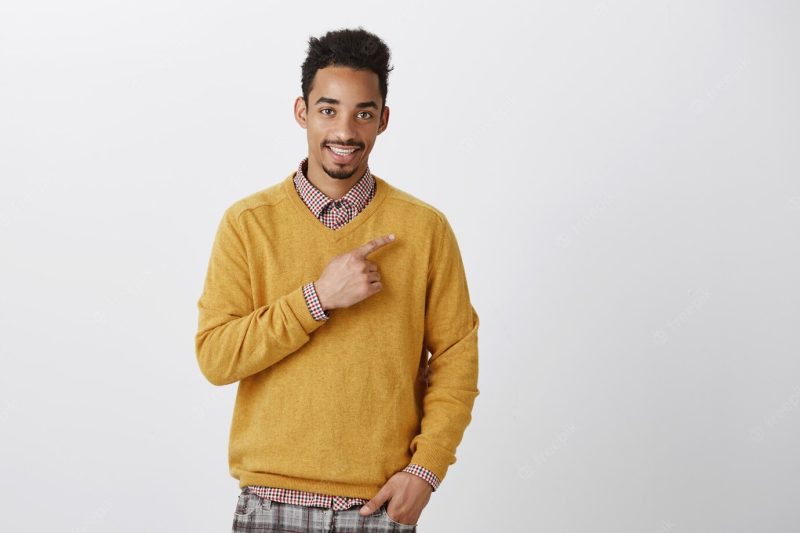 Time to visit new places. portrait of charming young dark-skinned guy with afro haircut smiling politely and pointing at upper right corner with index finger, smiling while promoting copy space Free Photo