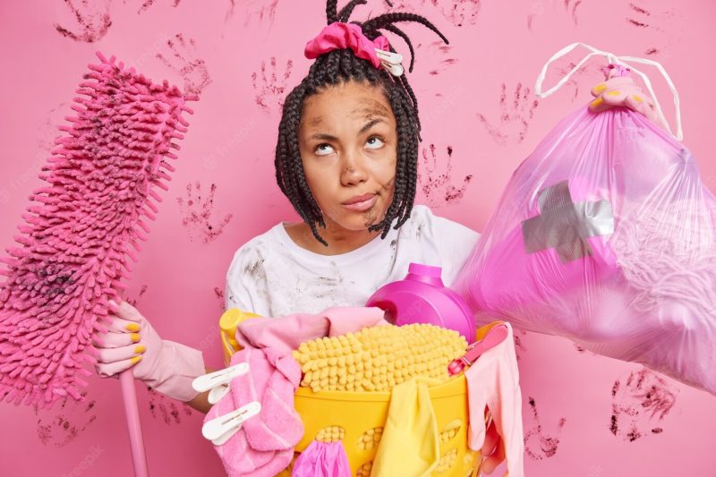 Tired thoughtful housewife poses with rubbish bag and mop stands dirty after doing housework cleans house uses cleaning products wears rubber protective gloves has dreadlocks isolated on pink wall Free Photo