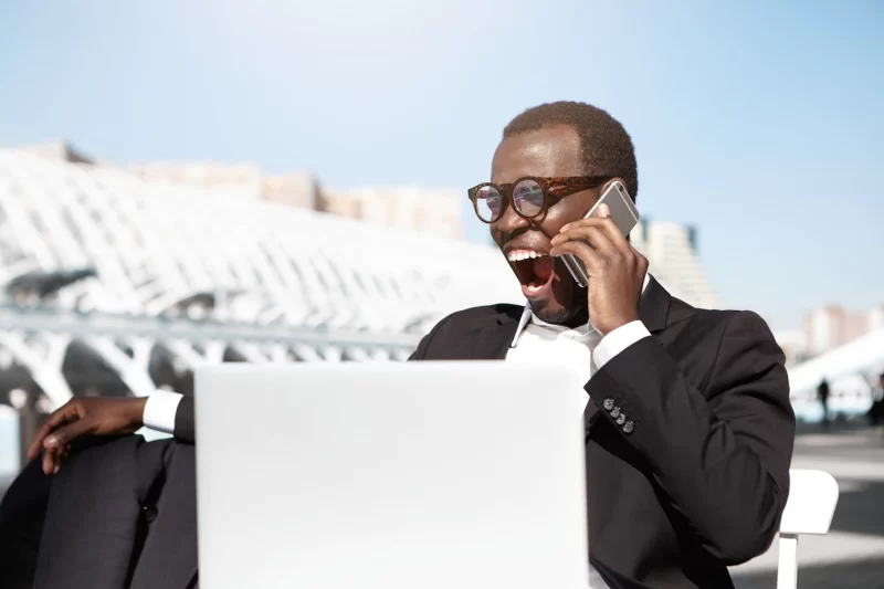 Tired yawning young Afro American banker dressed formally sitting at outdoor cafe table in front of notebook computer and talking on cell phone while waiting for his lunch, having bored sleepy look Free Photo