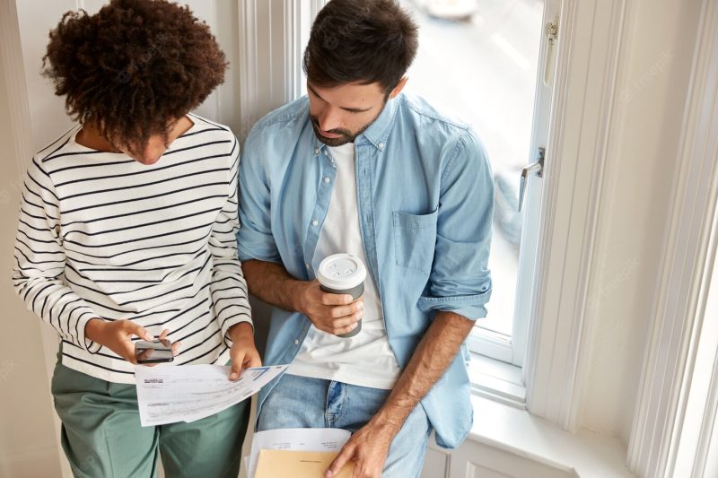 Top view of multi ethnic managers discuss marketing report, work together, check information on mobile phone drink takeaway coffee stand against window wall. unshaven man and his assistant Free Photo