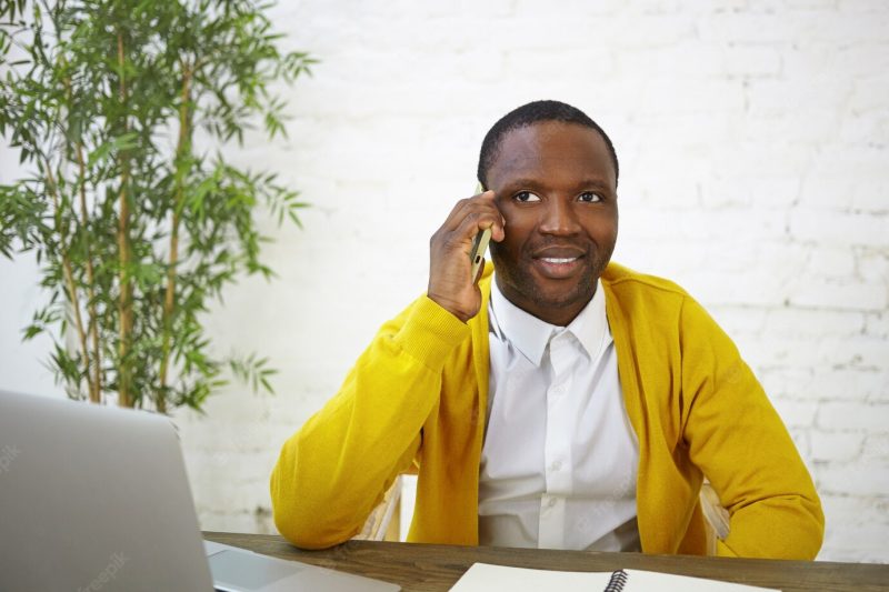 Trendy looking dark skinned male blogger having phone conversation, sitting in front of open laptop, working on content for his travel blog. people, job, occupation and modern electronic gadgets Free Photo