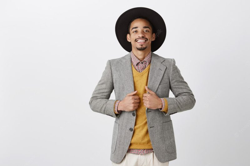 Trying new style, purchasing trendy jacket in store. portrait of happy successful dark-skinned bearded man in black hat, standing pleased and confident over gray wall, smiling with satisfaction Free Photo