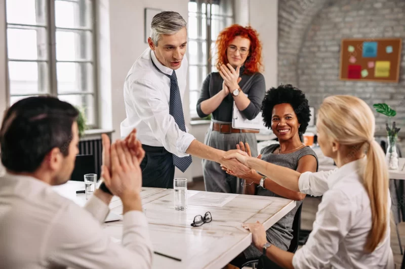 Two happy business people shaking hands on a meeting while other colleagues are applauding them in the office Free Photo