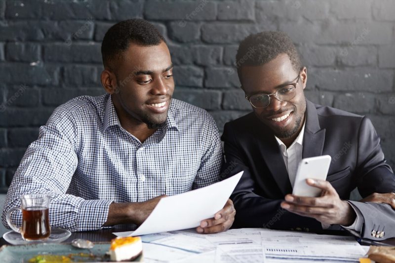 Two happy confident dark-skinned colleagues surfing internet on mobile phone while having break Free Photo