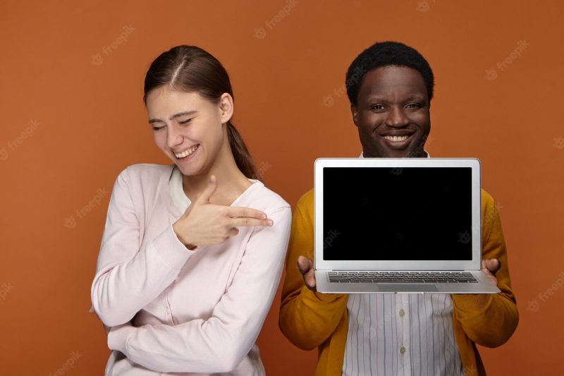 Two talented young professionals enjoying working together in team: cheerful confident black man holding generic laptop while positive cute white woman pointing at blank screen, showing presentation Free Photo