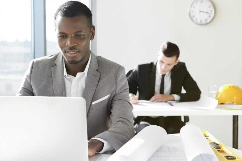 Two young architects of engineering company working in office. african designer developing new construction project using laptop, sitting at desk with rolls and ruler. Free Photo