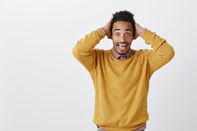 Unexpected good news make day bright. amazed good-looking african guy in casual yellow pullover holding hands on head from disbelief, being happy after positive event, standing over gray wall Free Photo