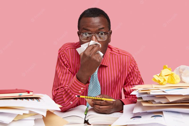 Unhealthy masculine adult uses handkerchief, has cold, running nose, works freelancer at home, carries spiral notebook Free Photo