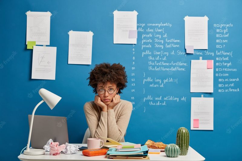 Upset dark skinned worker holds chin, sits at desk near laptop computer analyzes results feels tired of working. Free Photo