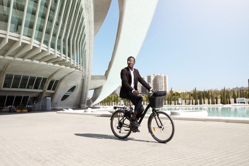 Urban lifestyle, ecology and transportation concept. fashionable modern ecologically friendly young afro american businessman wearing trendy round shades and formal suit cycling to work on bicycle Free Photo