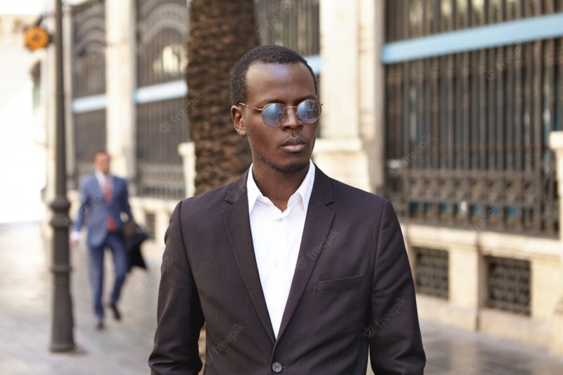 Urban outdoor portrait of confident serious young dark-skinned entrepreneur wearing stylish round shades and formal suit standing on street against office building Free Photo