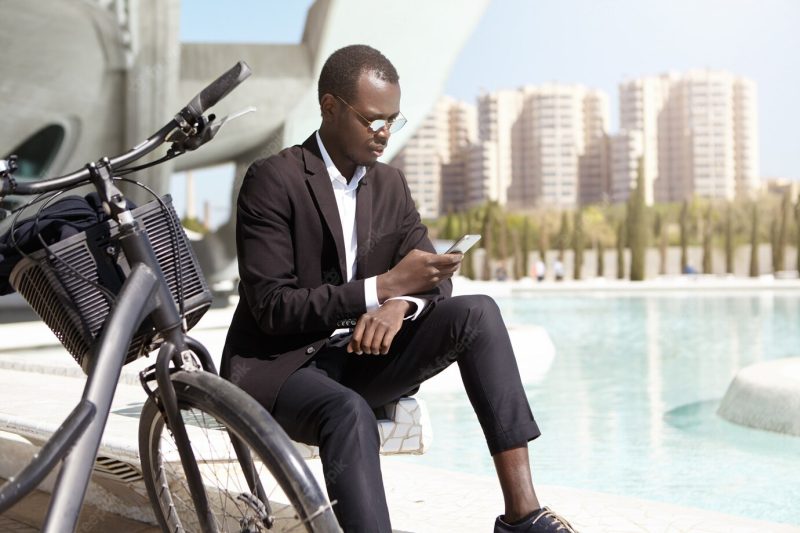 Urban shot of confident African American businessman wearing round sunglasses and elegant black suit sitting outdoors with his bicycle, using mobile phone, checking e-mail and dealing business issues Free Photo