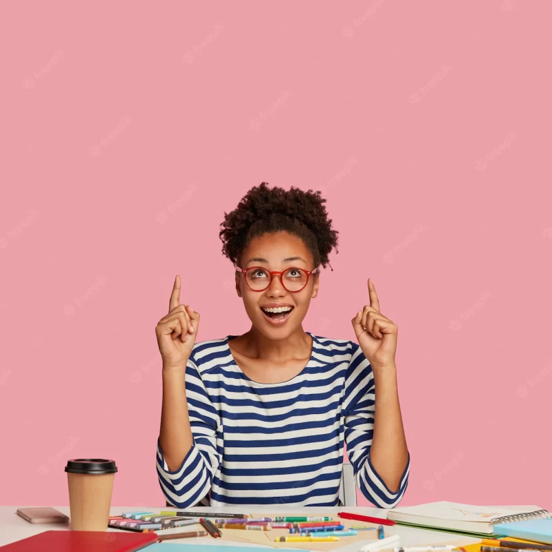 Vertical shot of glad dark skinned lovely woman artist, points with both index fingers, wears striped sweater, models against pink wall, feels excited, poses at desktop with spiral notebook Free Photo