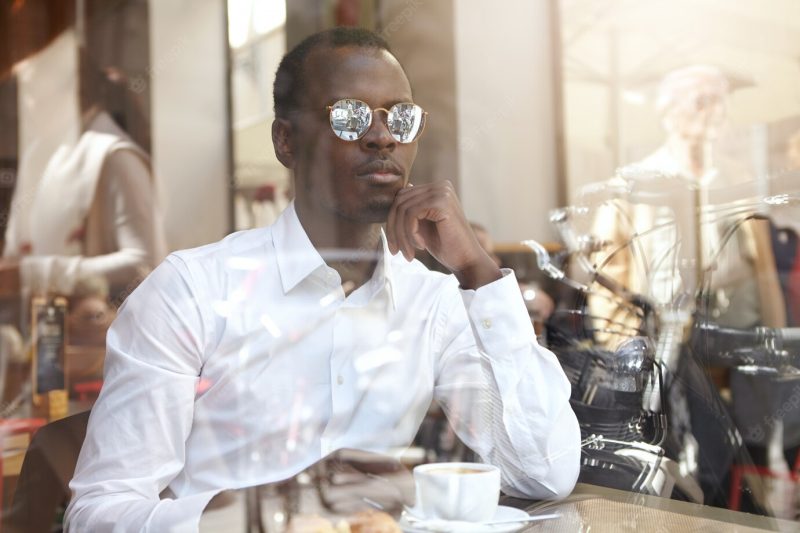 View from window glass of handsome pensive Afro American CEO in white shirt and stylish shades having black coffee during lunch, sitting alone at cafe, having thoughtful look, touching his chin Free Photo