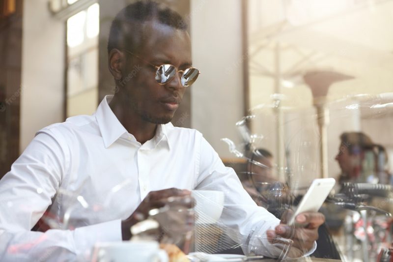 View through window glass of handsome black businessman or corporate worker wearing round shades and formal shirt drinking coffee and checking e-mail on mobile phone during break at modern cafe Free Photo