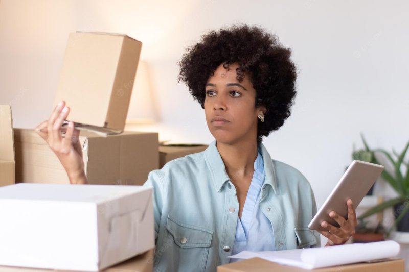 Warehouse clerk with afro hairstyle checking package. serious young woman using tablet while working among stacks of boxes. storehouse concept Free Photo