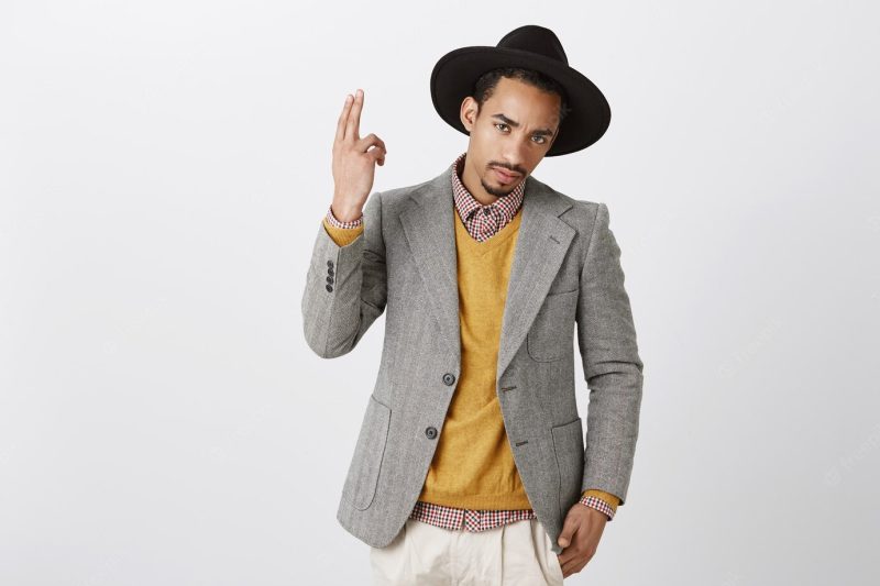 What are you looking at. confident good-looking dark-skinned man in black hat and gray jacket, showing gun gesture, lifting hand up Free Photo