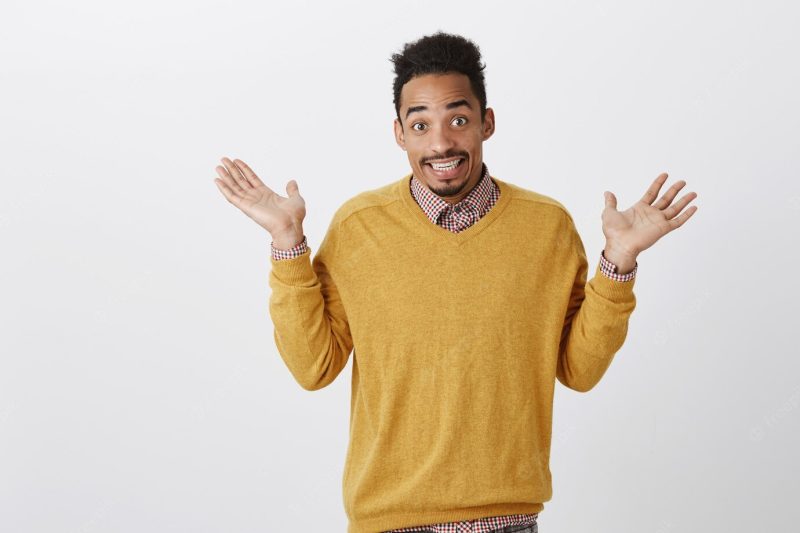 What done cannot be changed. portrait of confused unaware emotive dark-skinned guy with afro hairstyle raising palms in surrender, shrugging and smiling clueless, having no idea over gray wall Free Photo