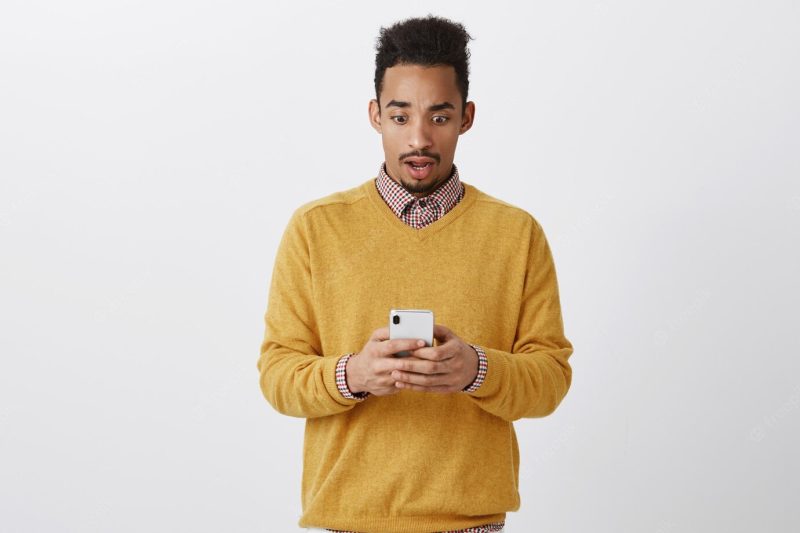 What the hell is going on. portrait of shocked and stunned attractive african-american, dropping jaw, being confused and surprised while reading article in internet via smartphone over gray wall Free Photo