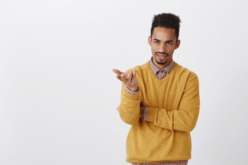 What is your point, dude. portrait of displeased dark-skinned male office worker in yellow pullover holding hands half-crossed, pointing aside with palm, expressing contempt and dislike, arguing Free Photo