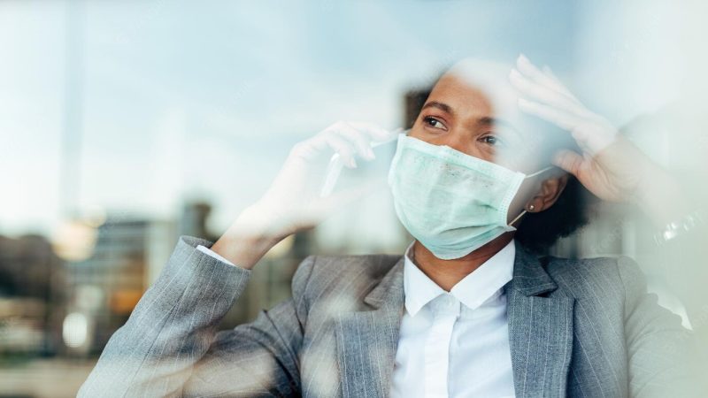 Worried african american businesswoman wearing protective mask and talking on mobile phone while looking through the window the view is through the glass Free Photo