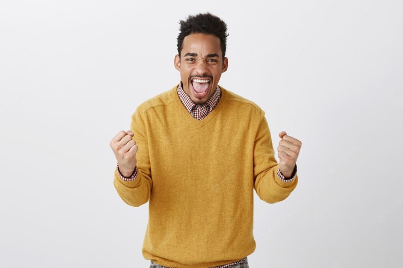 Yeah, man, we did it. portrait of excited good-looking dark-skinned male model shaking clenched fists and shouting from positive emotions, celebrating victory or cheering up favorite team Free Photo