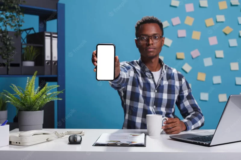 Young adult business person sitting in company modern workspace office while showing white screen isolated template to camera. project manager pointing phone device with mockup display to camera. Free Photo