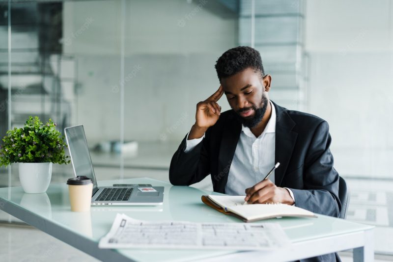 Young african businessman working in office at laptop and make notice in notebook Free Photo