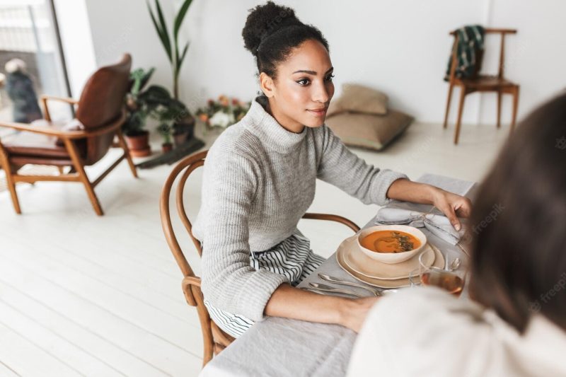 Young beautiful smiling african american woman with dark curly hair sitting at the table dreamily looking aside while spending time with friend in cozy cafe Free Photo