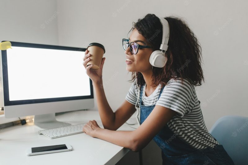 Young black woman wistfully looking away holding cup of coffee and smiling while working at office Free Photo