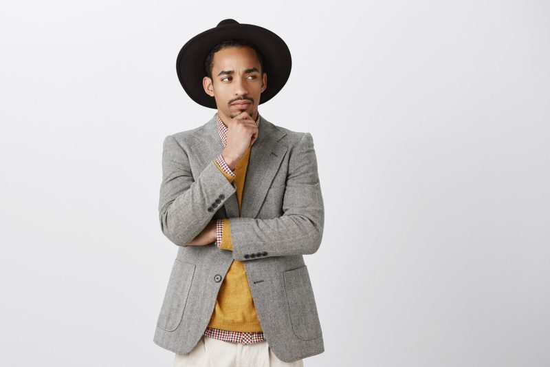 Young businessman doubting deal with foreign company. concerned good-looking dark-skinned guy with suspicious expression, standing in hat, touching chin and looking aside, thinking and focusing Free Photo