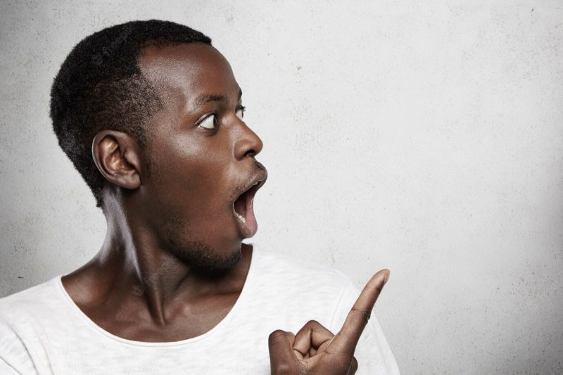 Young dark-skinned customer or employee looking in shock, opening mouth widely, showing something surprising pointing at white blank wall Free Photo