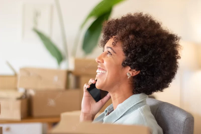 Young online shop worker calling client and smiling. happy black woman working in home office surrounded by cardboard parcels. startup business, e-commerce concept Free Photo