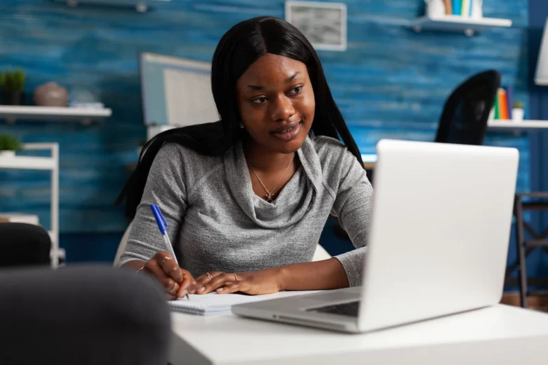 Young person taking notes on textbook paper with pen, looking at modern laptop. woman writing information on notebook files and doing remote work. adult working from home on business. Free Photo