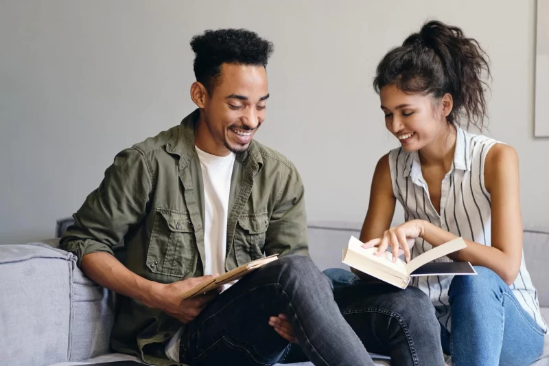 Young smiling African American man and pretty asian woman joyfully reading book together in modern co-working space Free Photo