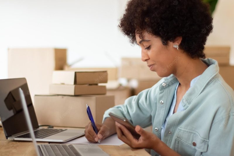 Young startup owner holding smartphone and checking order details. attractive black woman working on computer surrounded by cardboard parcels and making notes. business, e-commerce concept Free Photo