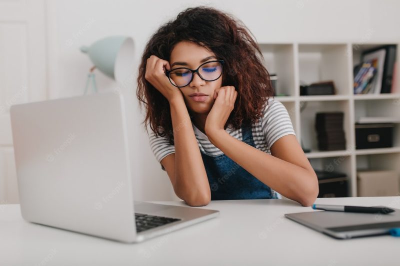 Young woman with light-brown skin wears trendy glasses sleeping in front of laptop in office Free Photo