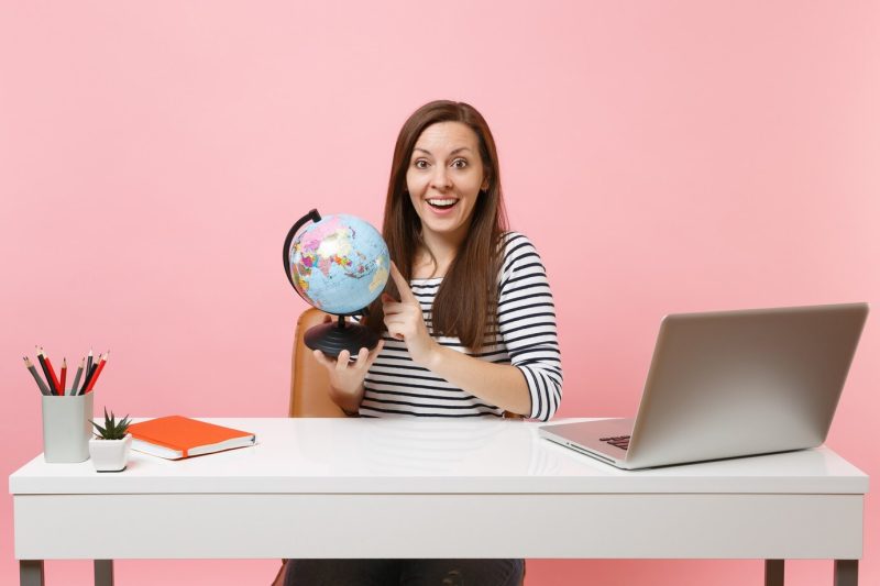 Amazed woman holding world globe and planning vacation while sit and work at white desk with contemporary pc laptop Free Photo