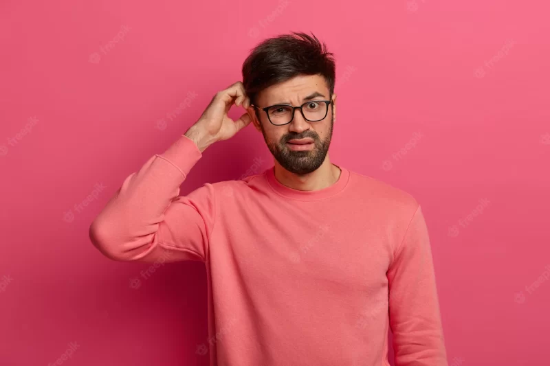 Serious bearded young man scratches head, thinks deeply about how to solve problem Free Photo