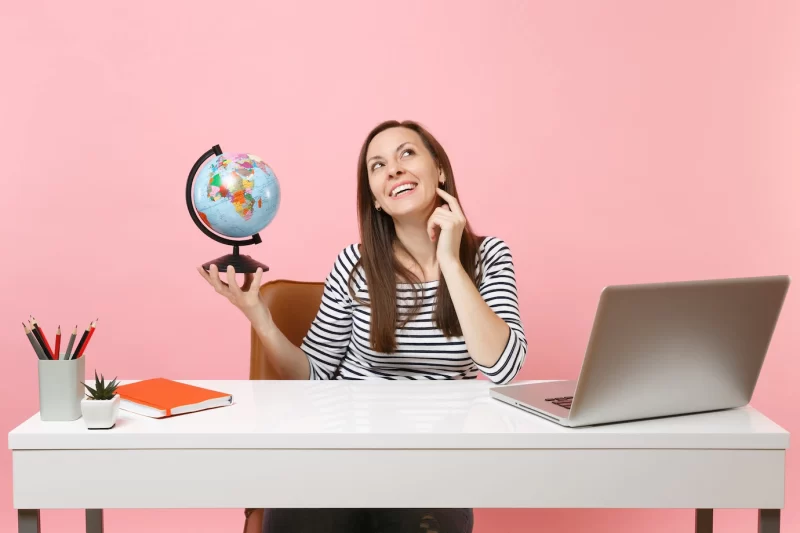 Young pensive woman holding globe, dreaming planning vacation while sit, work at office with contemporary pc laptop Free Photo