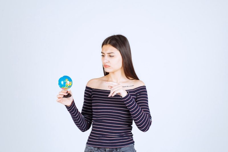 Young woman in striped shirt holding a mini globe and guessing a place over it Free Photo